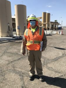 Engineer wearing coveralls, safety vest, hard hat, standing by water treatment system.